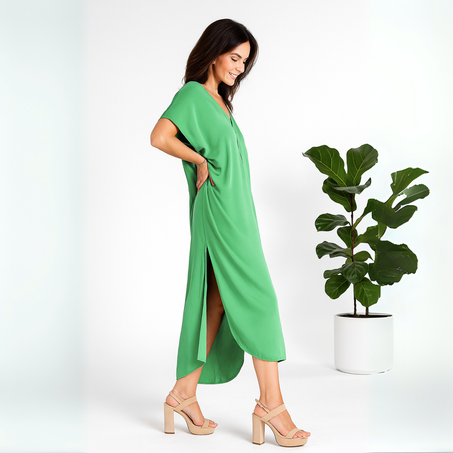 Side view of a woman wearing a green caftan walking in a white studio with a fig tree behind her.