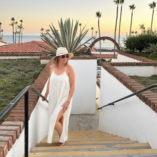 Woman in a white dress and hat standing on a staircase with palm trees and ocean in the background
