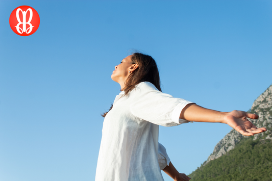 Woman outside with her eyes closed opening her arms up and facing up to the sun with a blue sky behind her.
