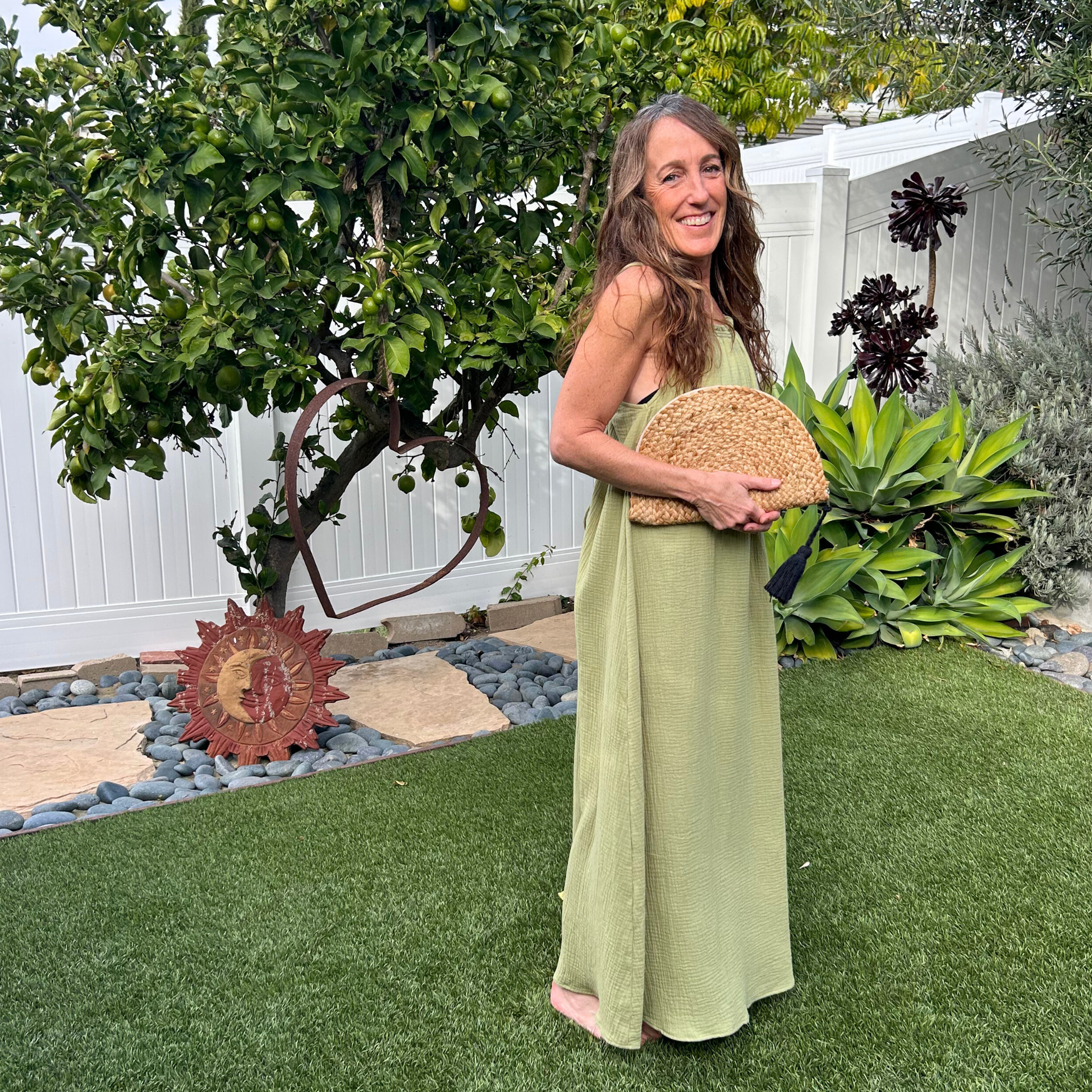 Woman standing in a yard wearing a green dress and carrying a woven clutch.