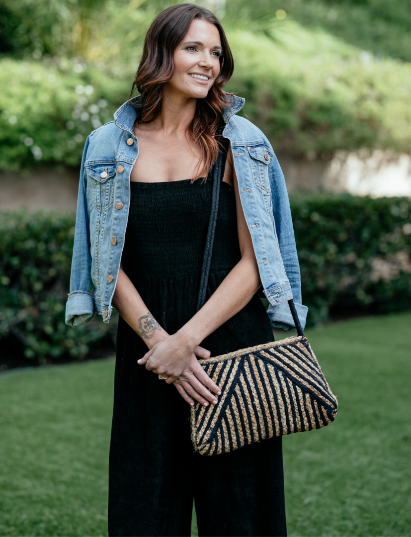 Woman wearing black jumpsuit paired with jean jacket and jute purse.