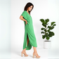Side view of a woman wearing a green caftan walking in a white studio with a fig tree behind her.