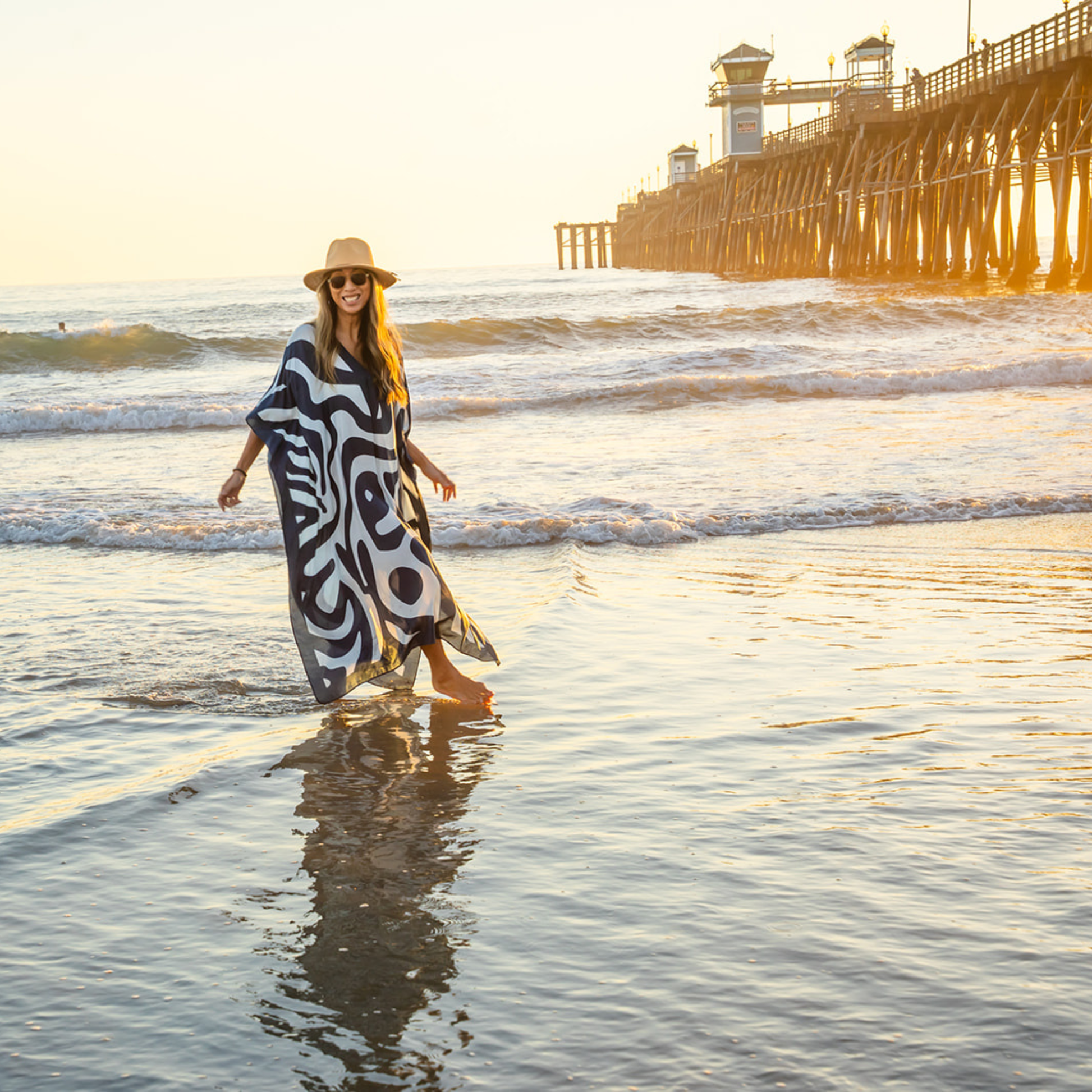 Woman in a blue and white patterned kaftan and hat walking on a beach with a pier in the background.