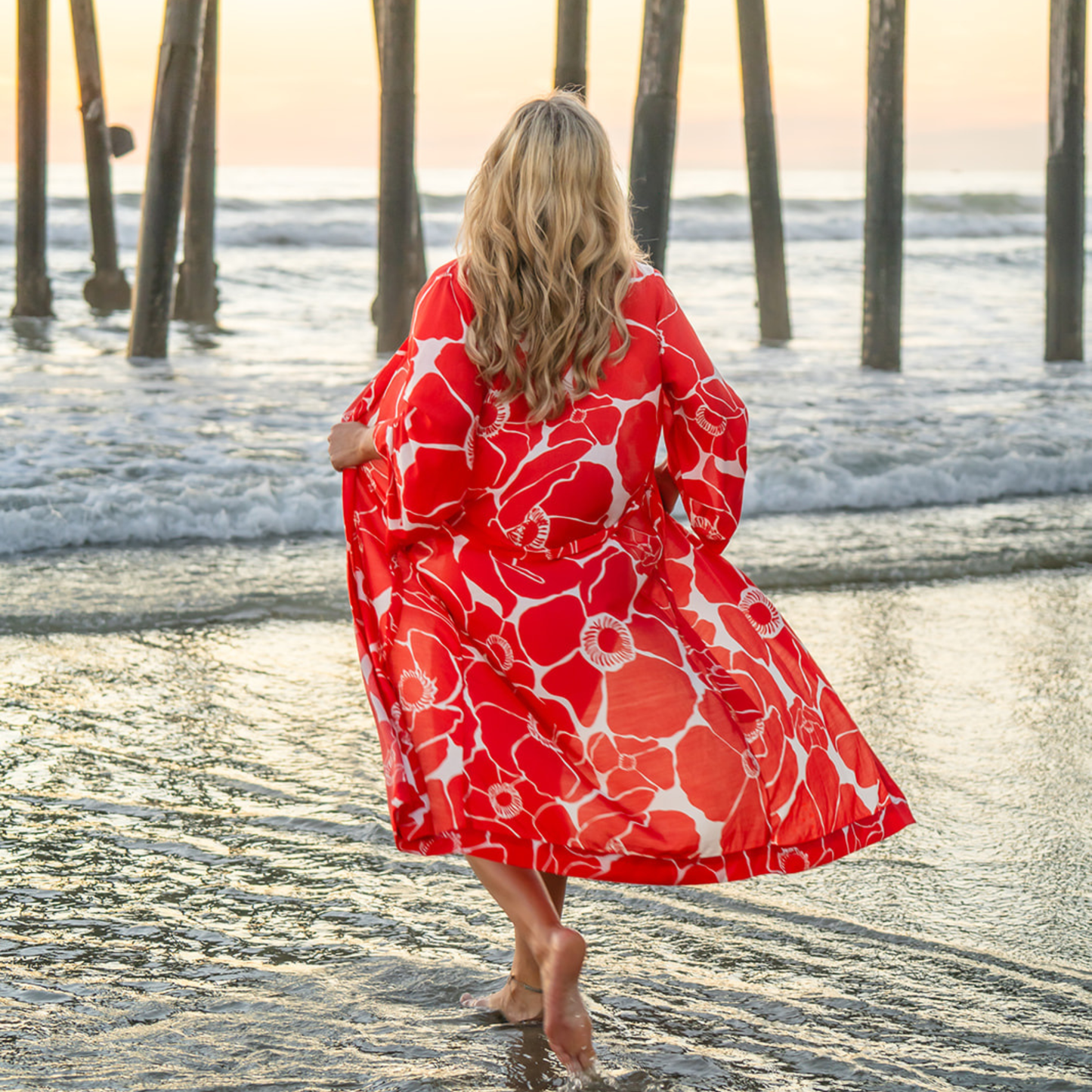 Woman in a red floral dress walking on a beach with wooden poles in the background.