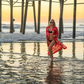 Woman in a red cover-up standing in shallow water near a pier at sunset.