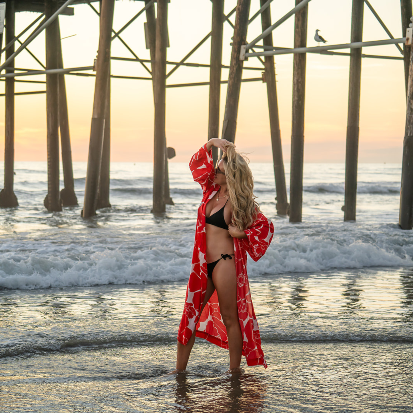 Woman in a red floral cover-up standing in the ocean under a pier at sunset.