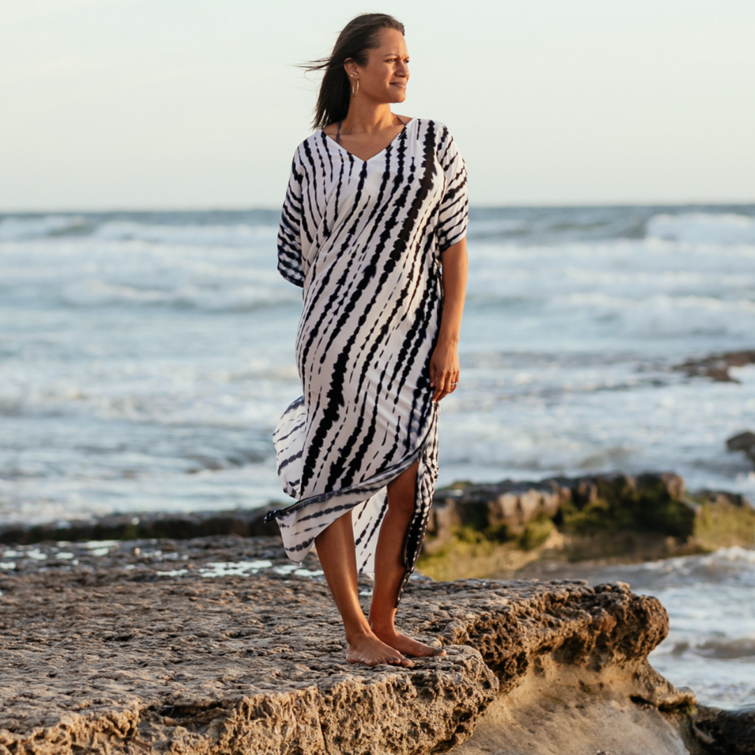 Woman standing at the ocean's edge at low tide looking at the sunset. She's wearing a tie dyed caftan.