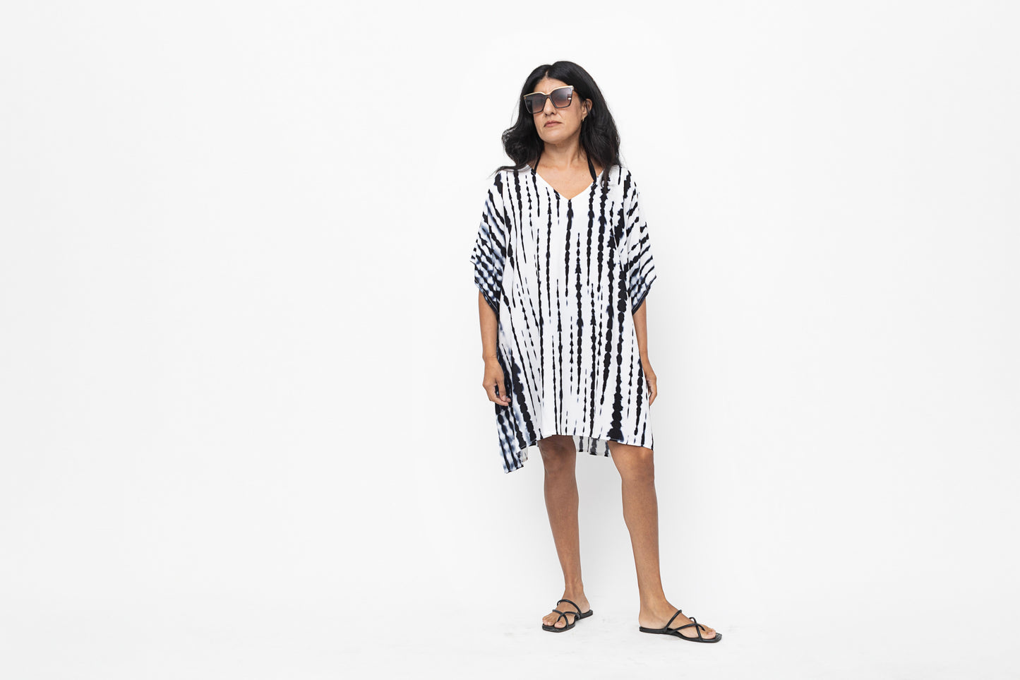 Woman standing in a blue and white short caftan wearing gold sandals and sunglasses.
