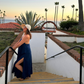 Woman in a blue lightweight linen dress and sun hat standing on a staircase with palm trees and ocean in the background.