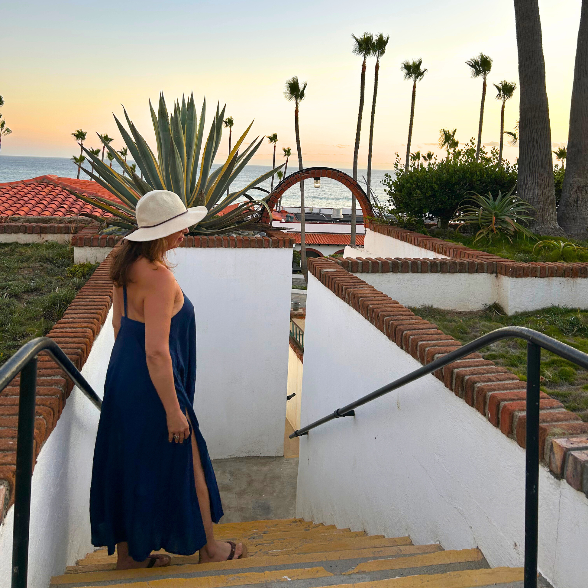 Woman in a blue dress and white hat standing on a staircase with palm trees and ocean in the background.