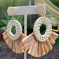 A pair of hoop earrings with natural raffia tassels in ivory and brown tones, displayed on a wooden stand.