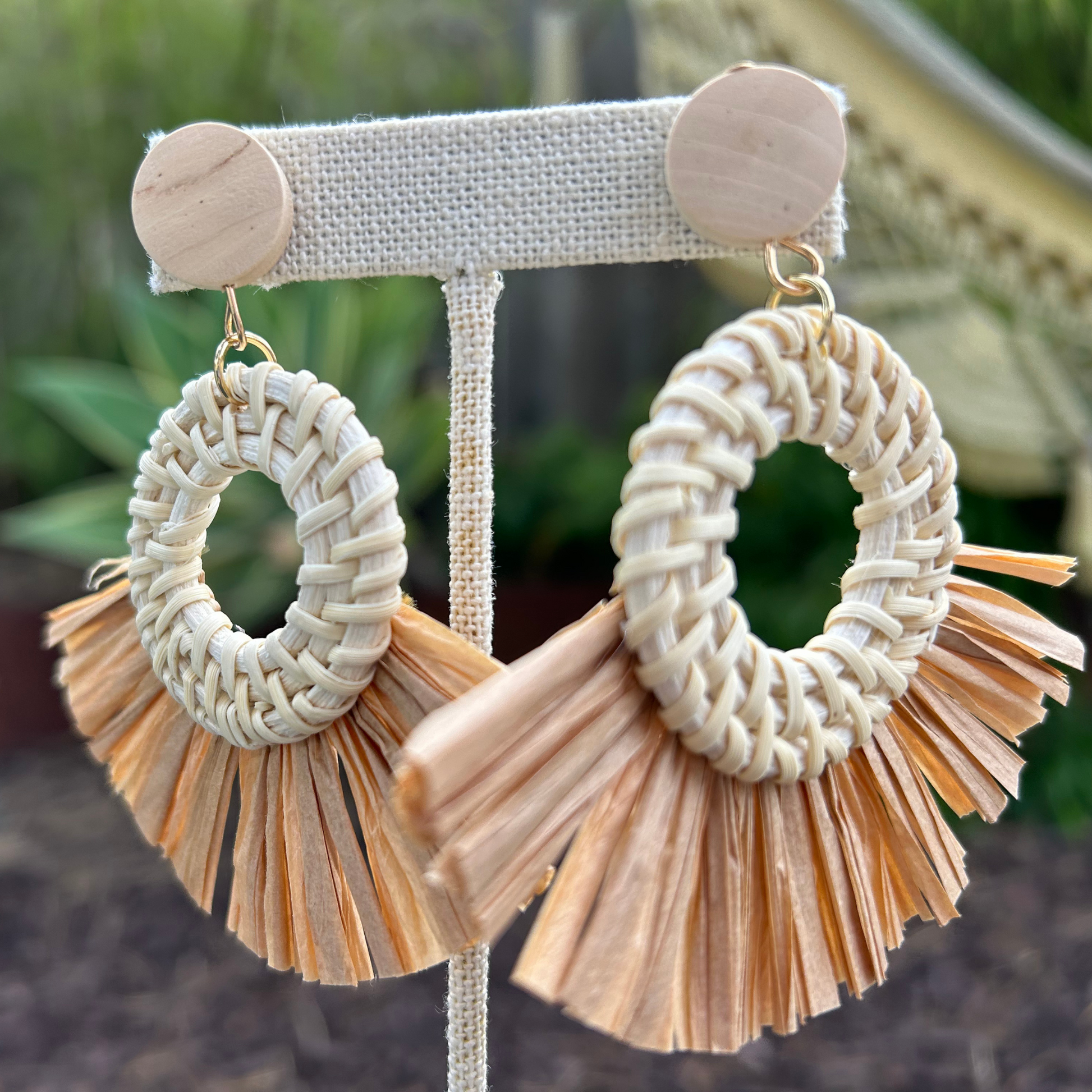A pair of hoop earrings with natural raffia tassels in ivory and brown tones, displayed on a wooden stand.