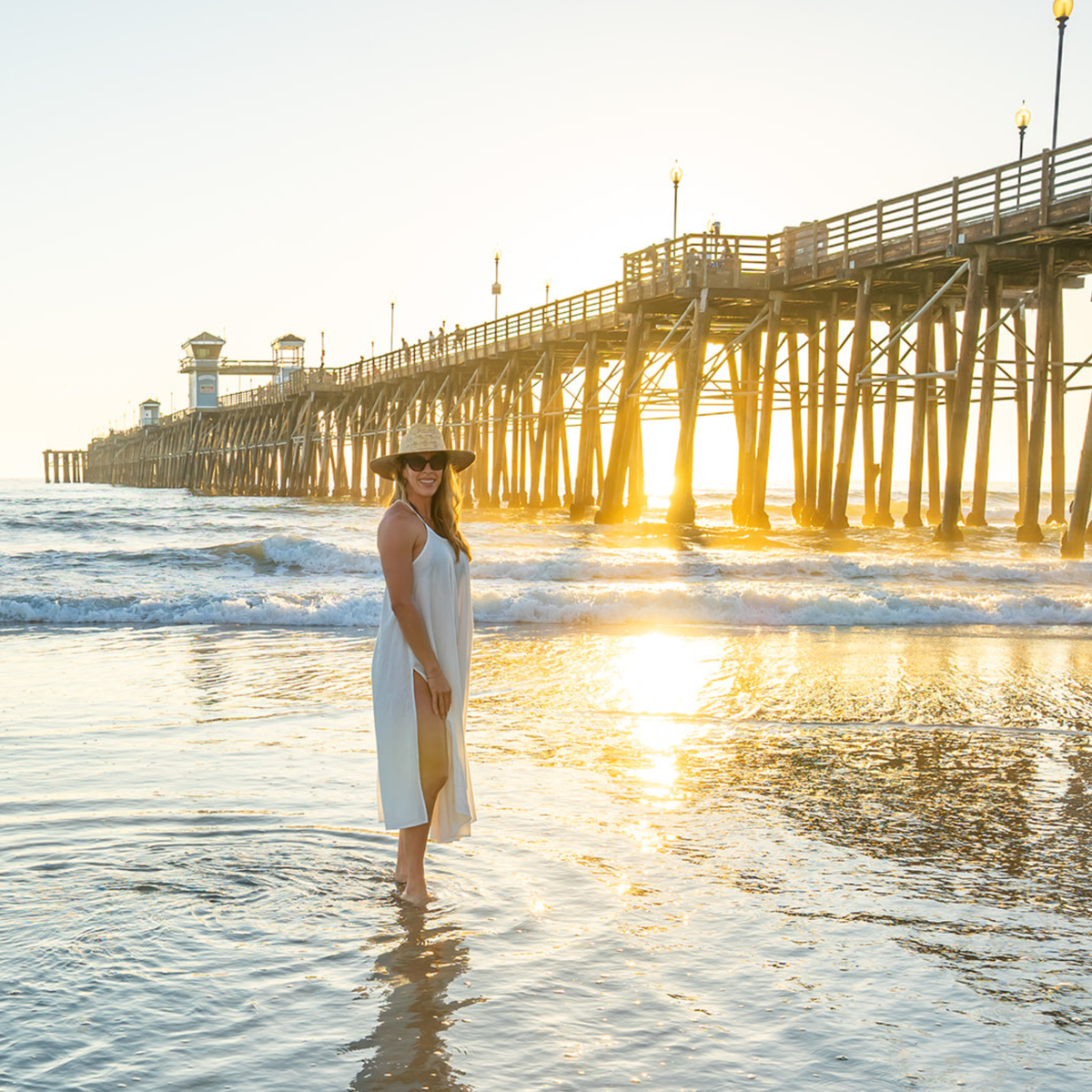 Woman in a white dress standing on a beach with a pier in the background during sunset.