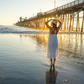 Woman in a white dress standing on a beach with a pier in the background during sunset.
