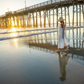Woman in a white dress and hat standing on a beach with a pier in the background during sunset.