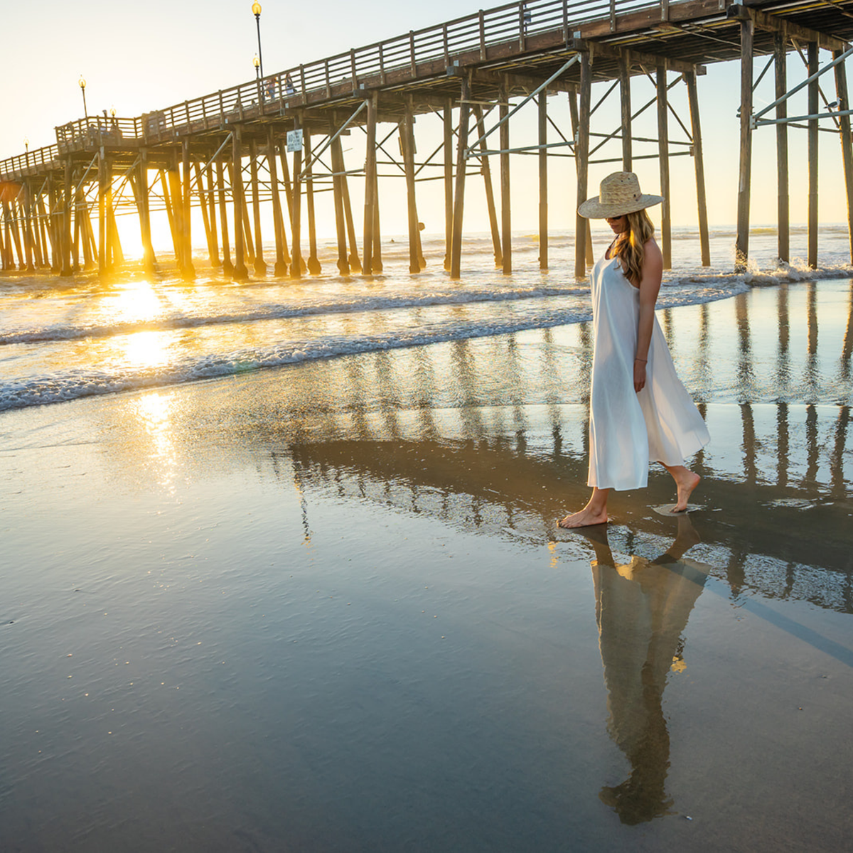 Woman in a white dress and hat standing on a beach with a pier in the background during sunset.