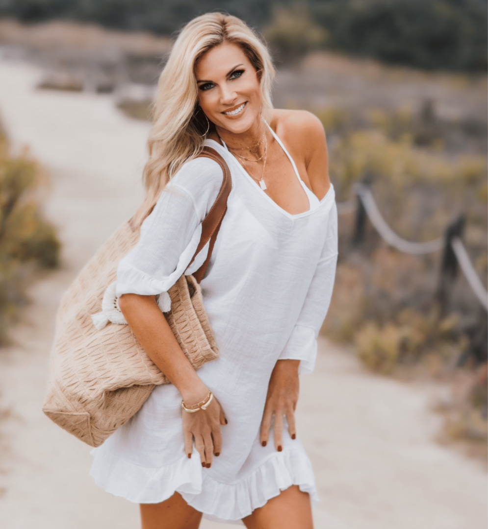 Woman posing in white cover up and beach bag in a sandy pathway.