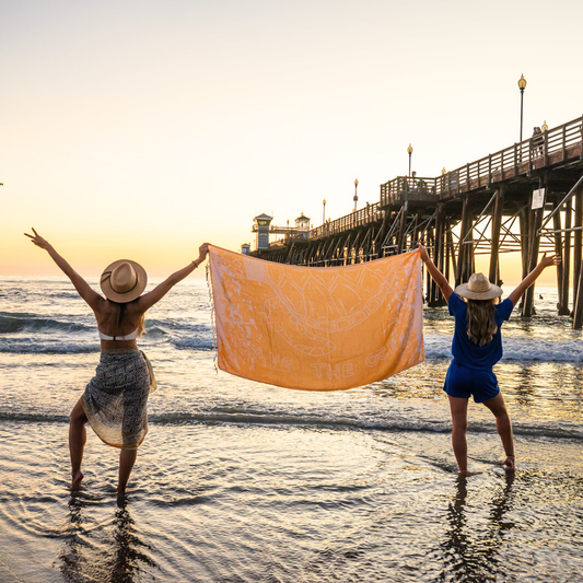 Two people on a beach holding a large yellow towel that says "Save The Ocean" with a sunset and pier in the background.