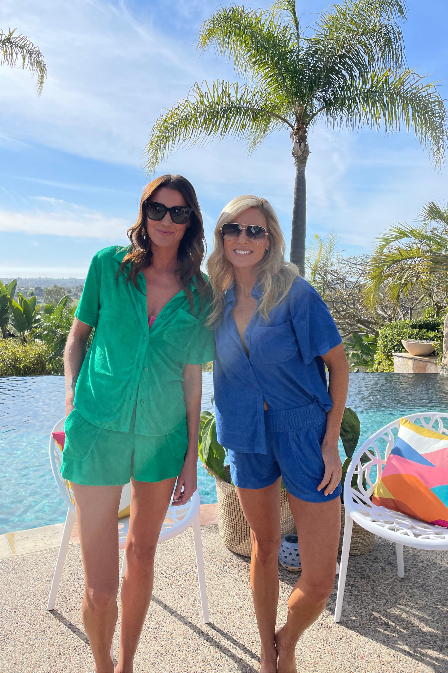 Two women posing in a green and a blue matching shorts set standing in front of a tropical pool.