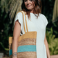 Woman in white dress carrying straw bag with yellow and turquoise stripes, looking down to the side.