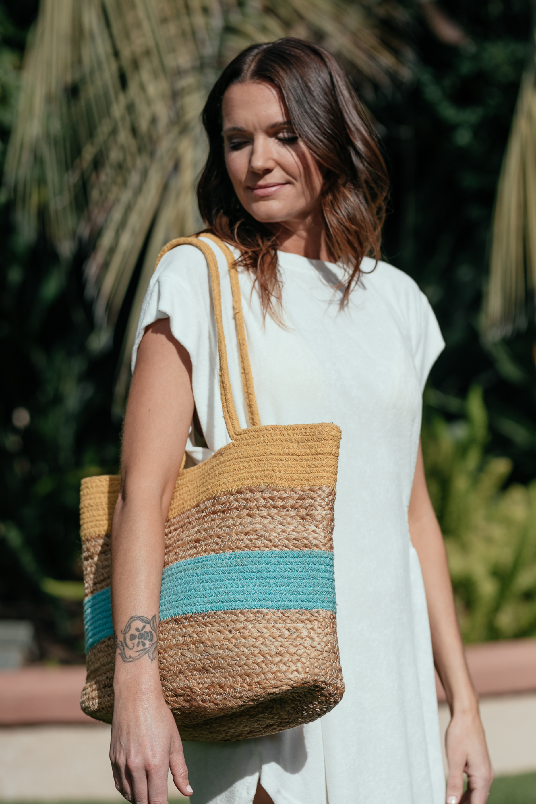 Woman in white dress carrying straw bag with yellow and turquoise stripes, looking down to the side.