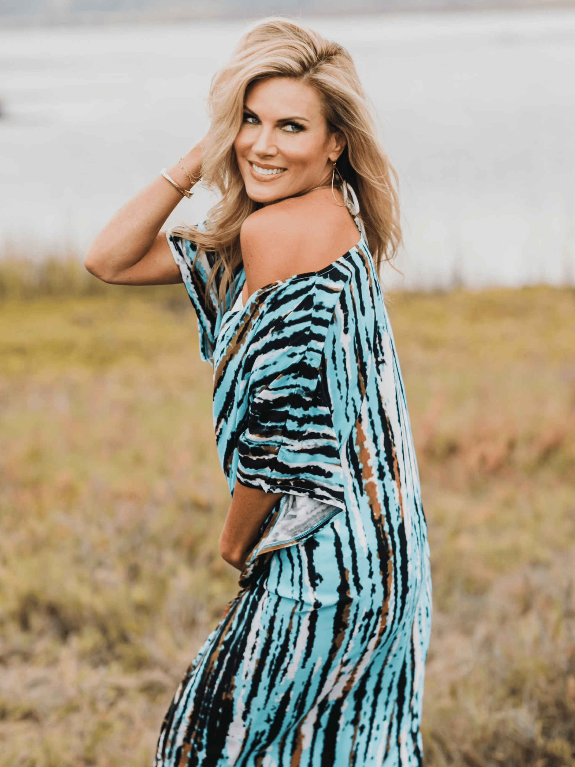 Woman in tie dyed caftan turned sideways towards the camera, looking over her shoulder. She is in a field.