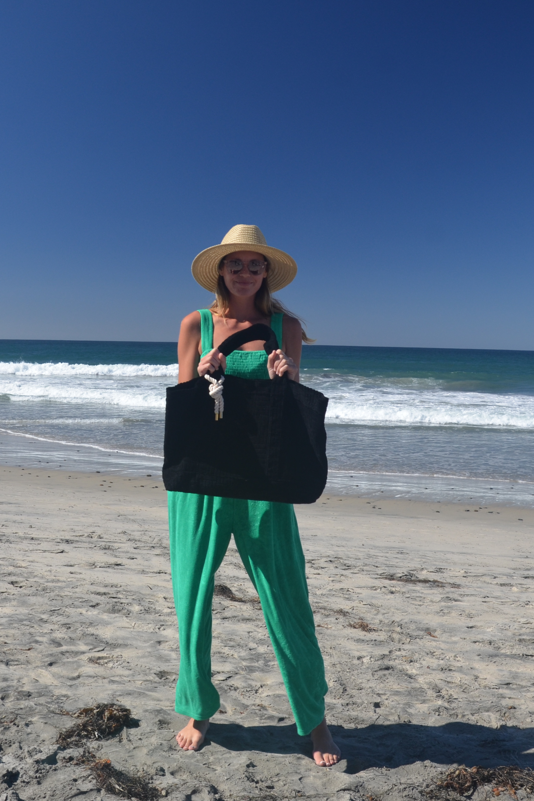 Woman on a beach wearing a green jumpsuit, holding a black bag.